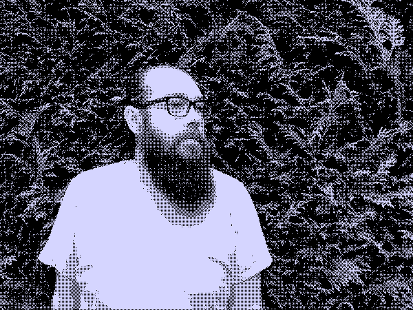 A person with a beard to their shoulders, wearing thick glasses and a t-shirt, standing in front of a hedge.